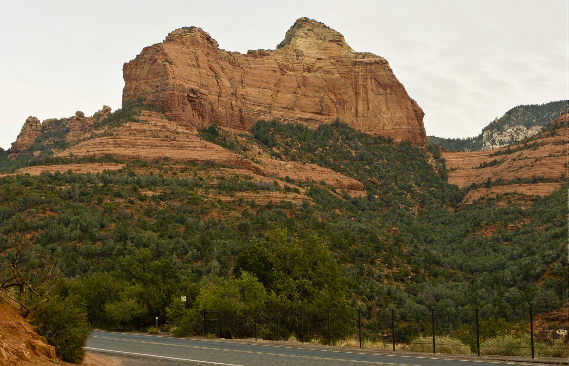 Large red rock formation with green trees, road in foreground, under overcast sky.