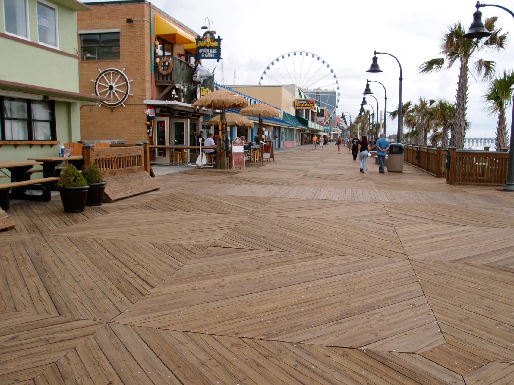 Wide wooden boardwalk with shops, palm trees, and a distant Ferris wheel.
