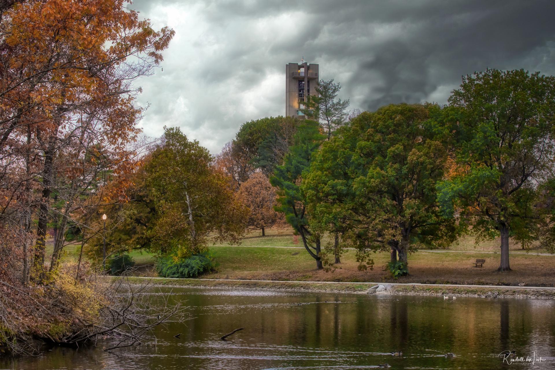Overcast day over a pond with colorful autumn trees and a distant tower.