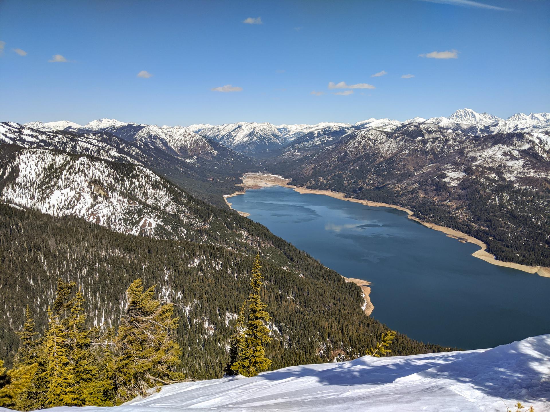 Sunny mountain vista with a vast lake, snow, and dense pine trees.