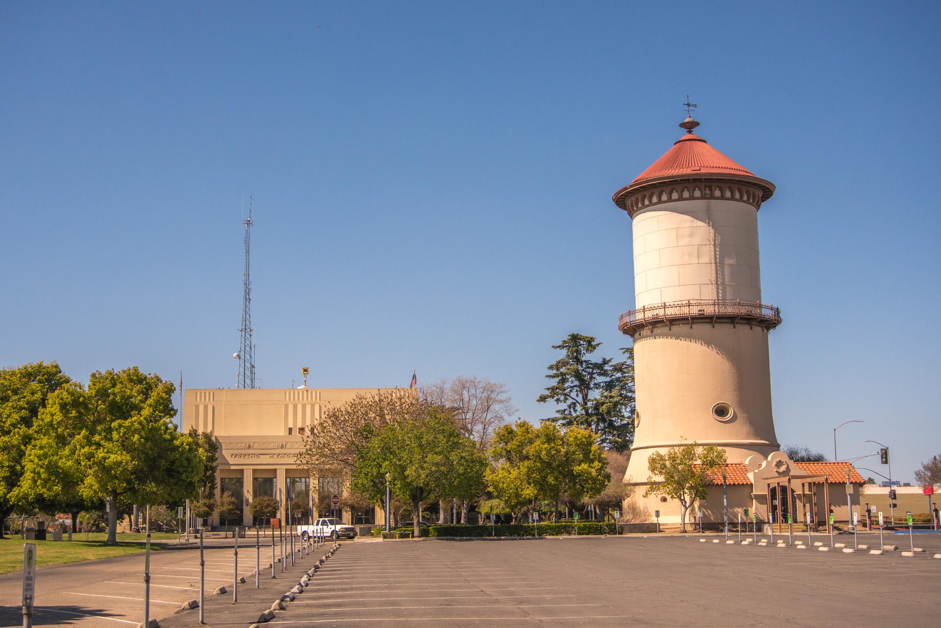 Tall water tower with a red roof, large building, and trees under clear sky.