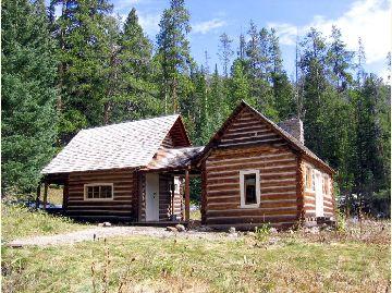 Two rustic log cabins in a forest clearing on a sunny day.