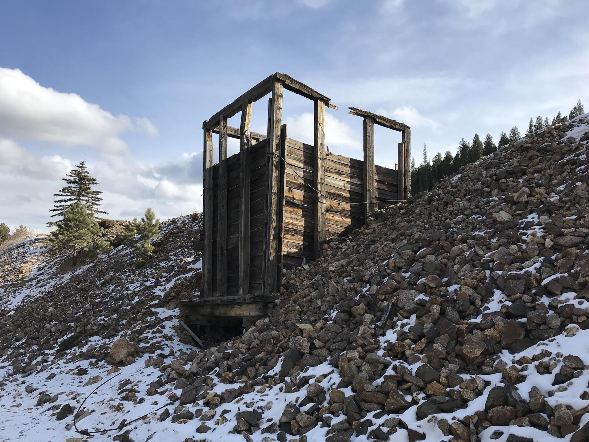Dilapidated wooden structure on a rocky, snowy hillside under a bright sky.