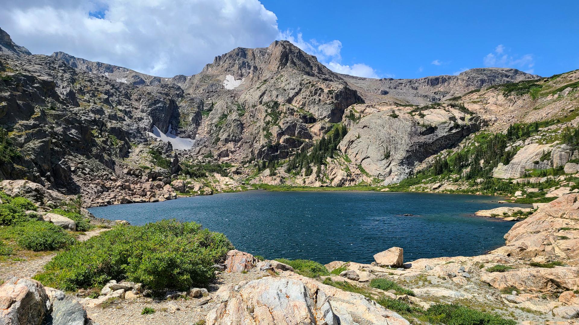 Serene mountain lake surrounded by rocky peaks and green shrubs under blue sky.