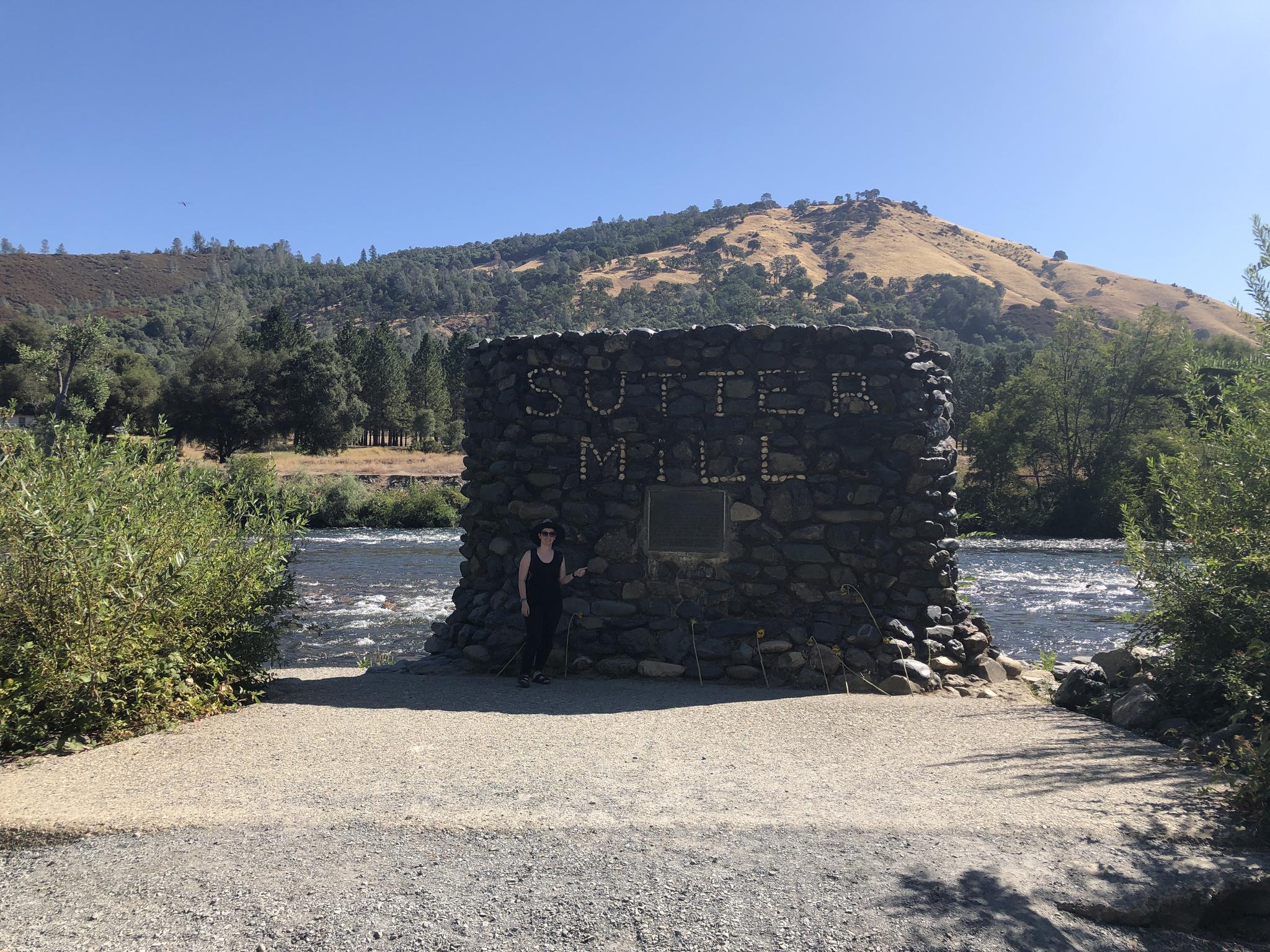 Woman stands by a stone monument next to a river and sunny hills.