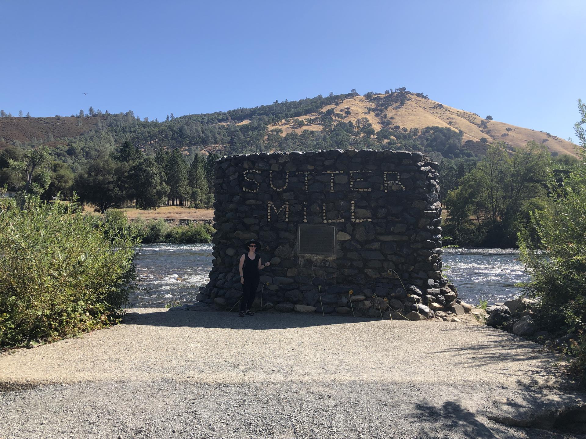 Woman stands by a stone monument next to a river and sunny hills.