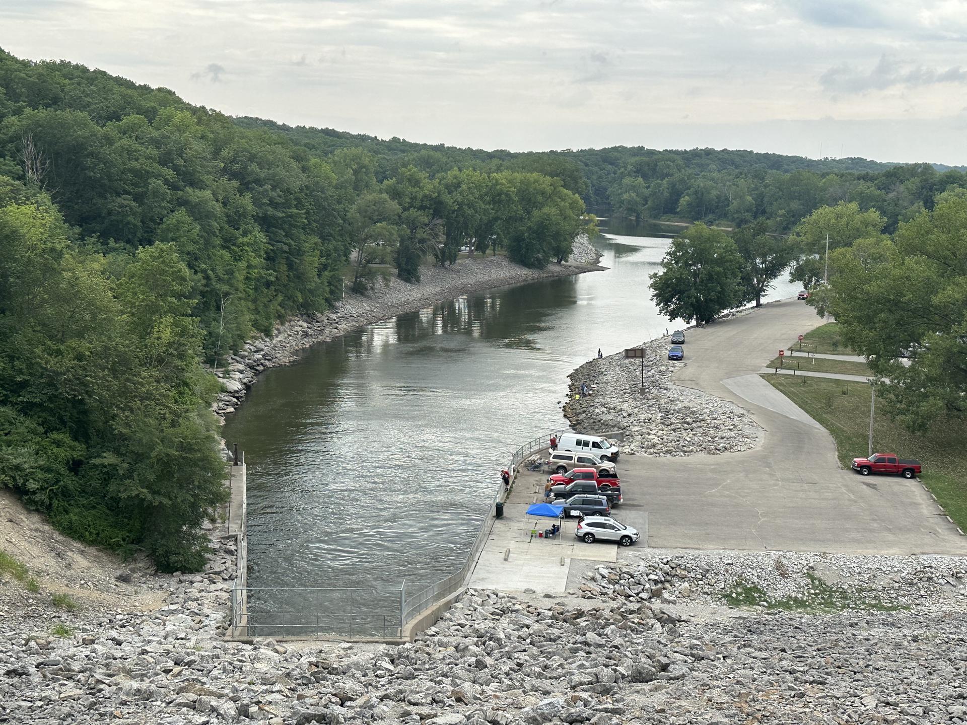 Overcast view of a river, boat ramp, cars, and people on rocky banks.