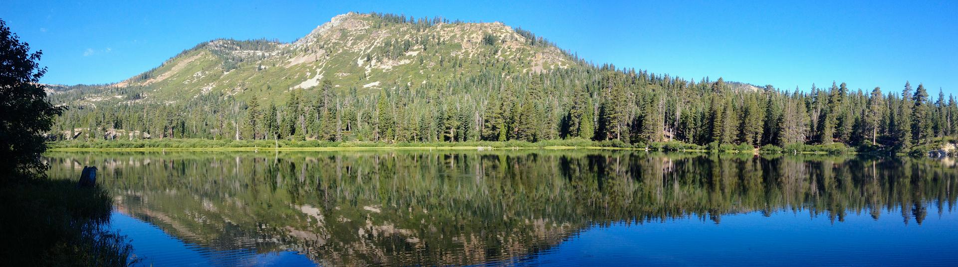 Panoramic view of still lake reflecting forested mountain under clear blue sky.