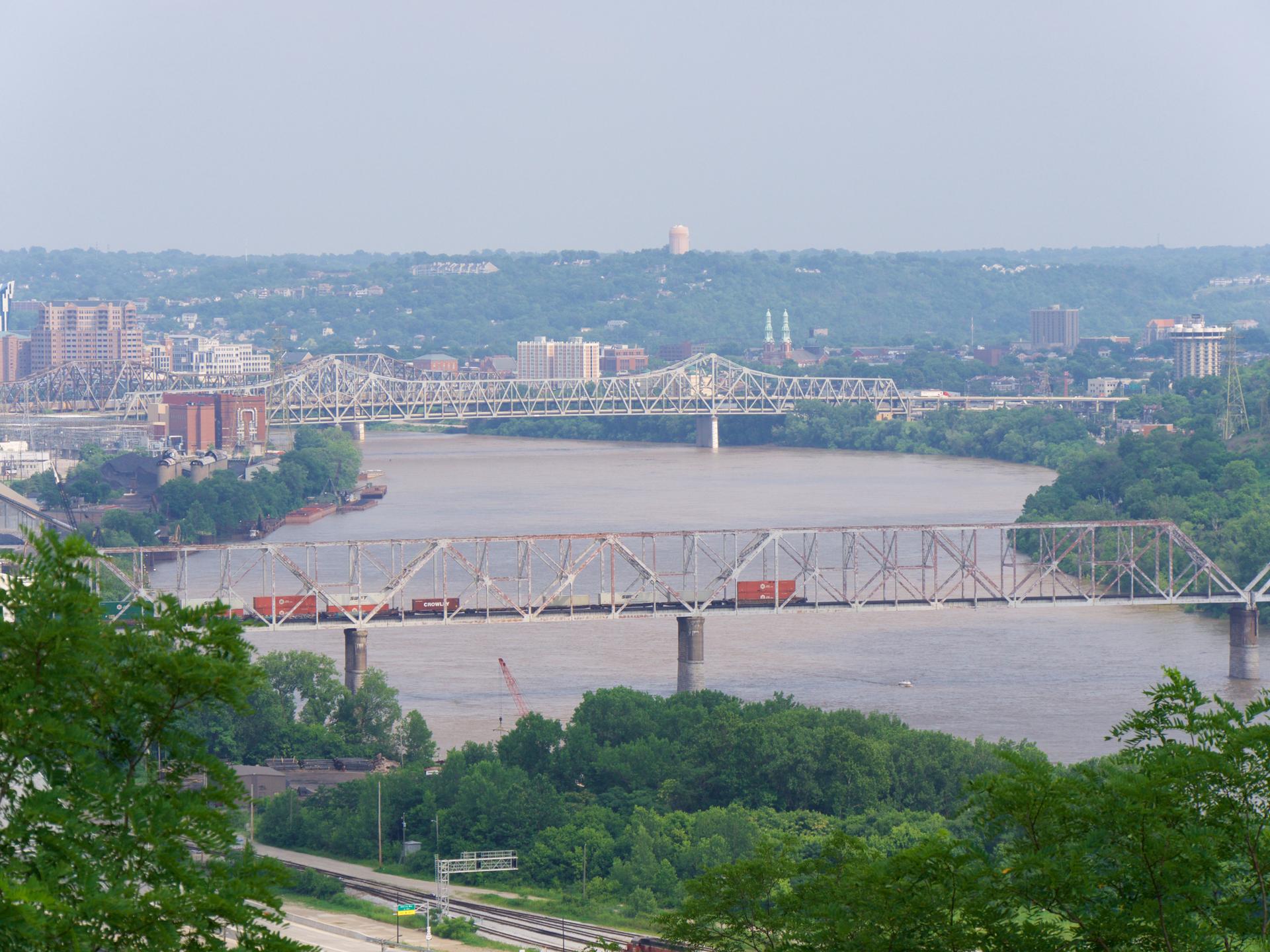 Multiple large bridges span a wide river with a train and distant city skyline.