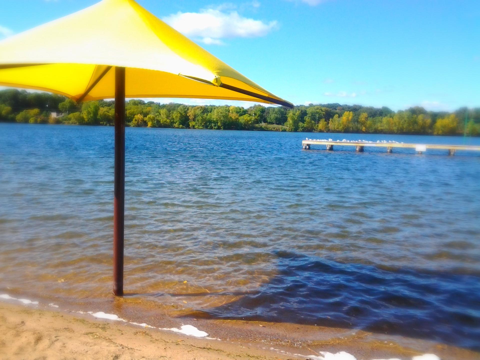 Yellow umbrella on a sandy beach next to a lake with a distant dock.