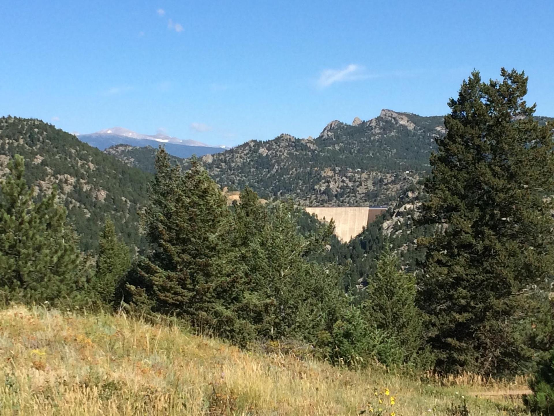 A large dam is visible in a valley surrounded by forested mountains under a clear sky.