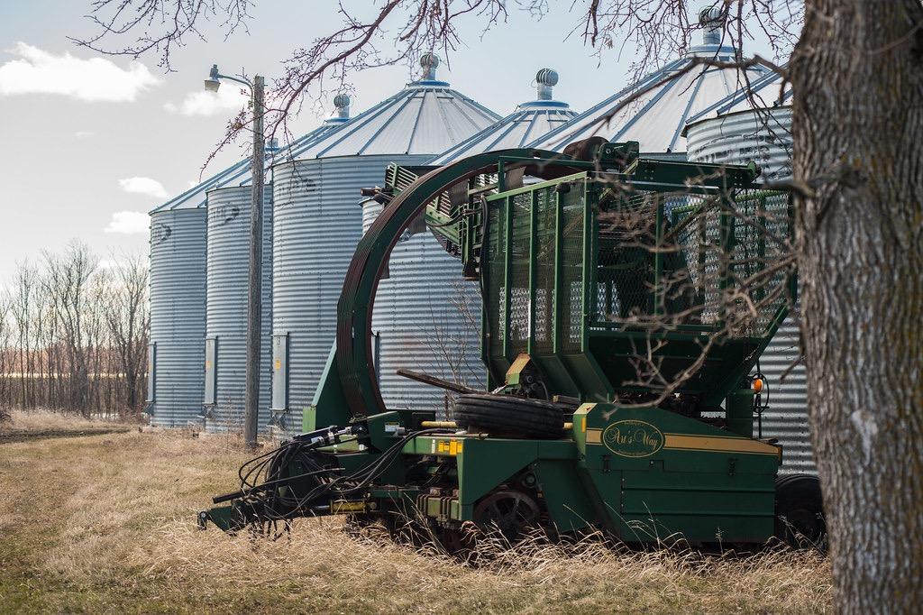 Green farm machinery sits in dry grass before silver silos and bare trees.