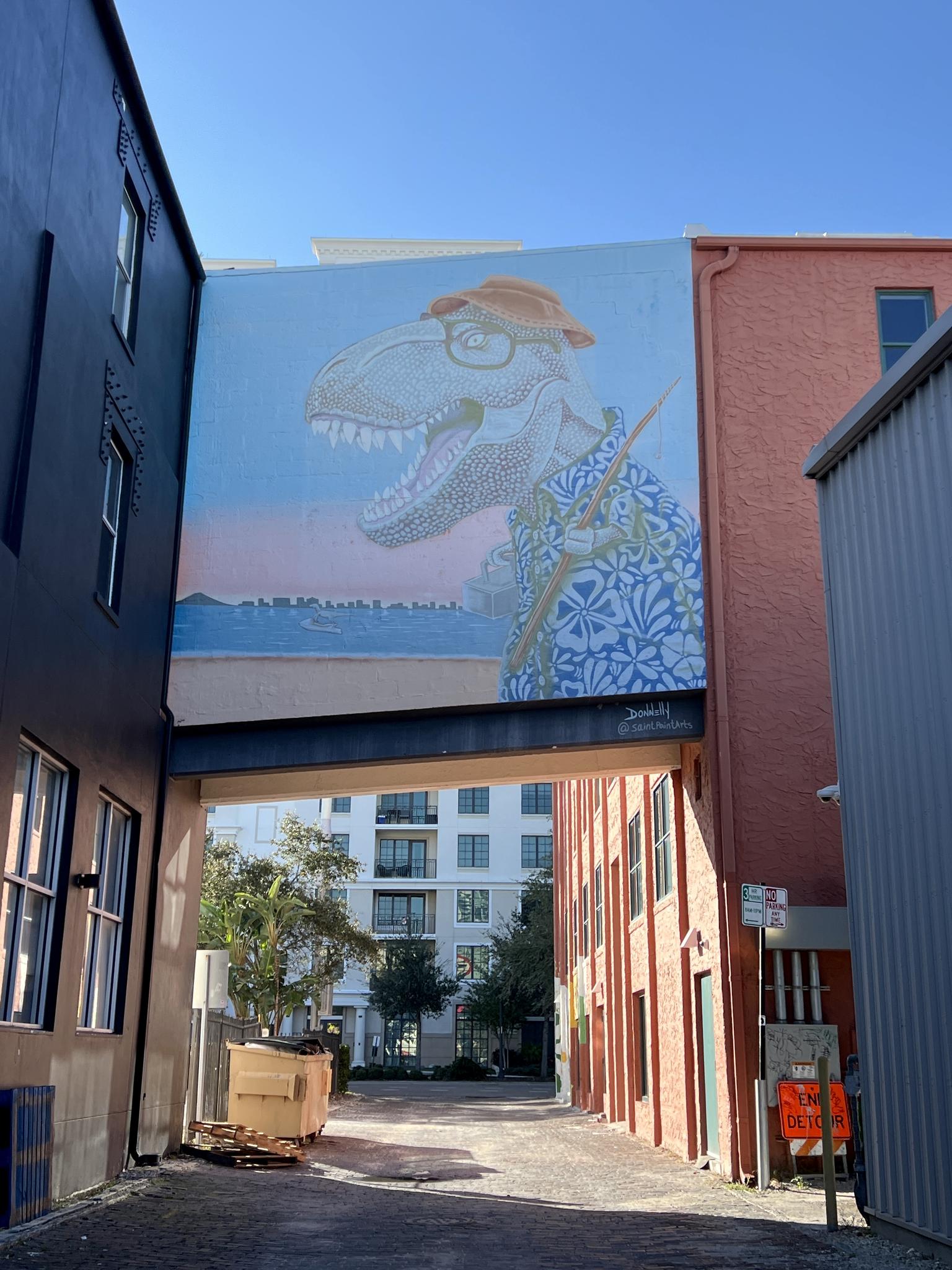 A large mural of a dinosaur wearing glasses and a shirt in an urban alleyway.