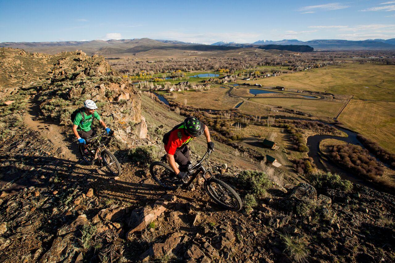 Two mountain bikers on a rocky ridge overlooking a valley at sunset.