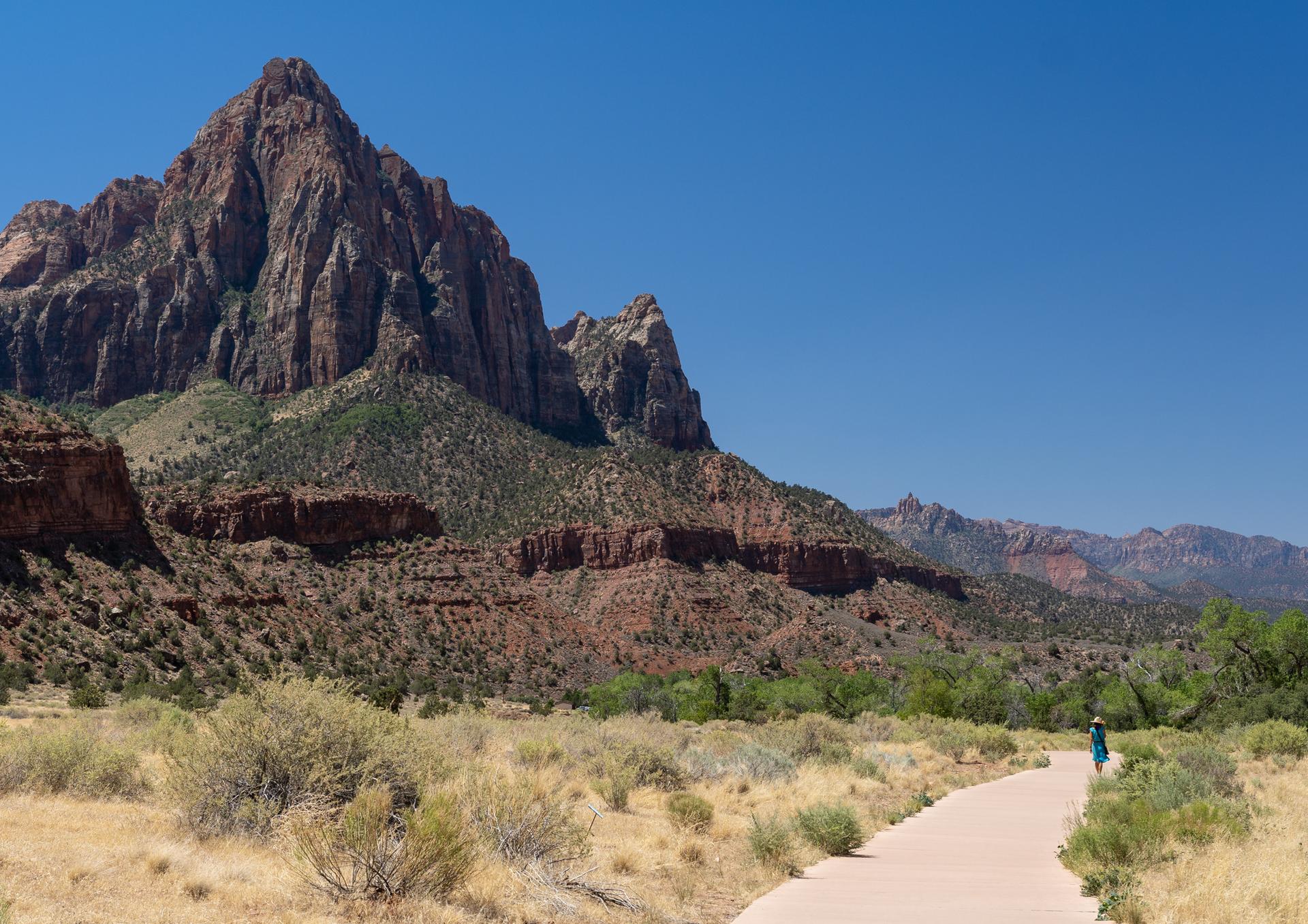 A person walks on a paved path towards a towering red rock mountain under a clear sky.