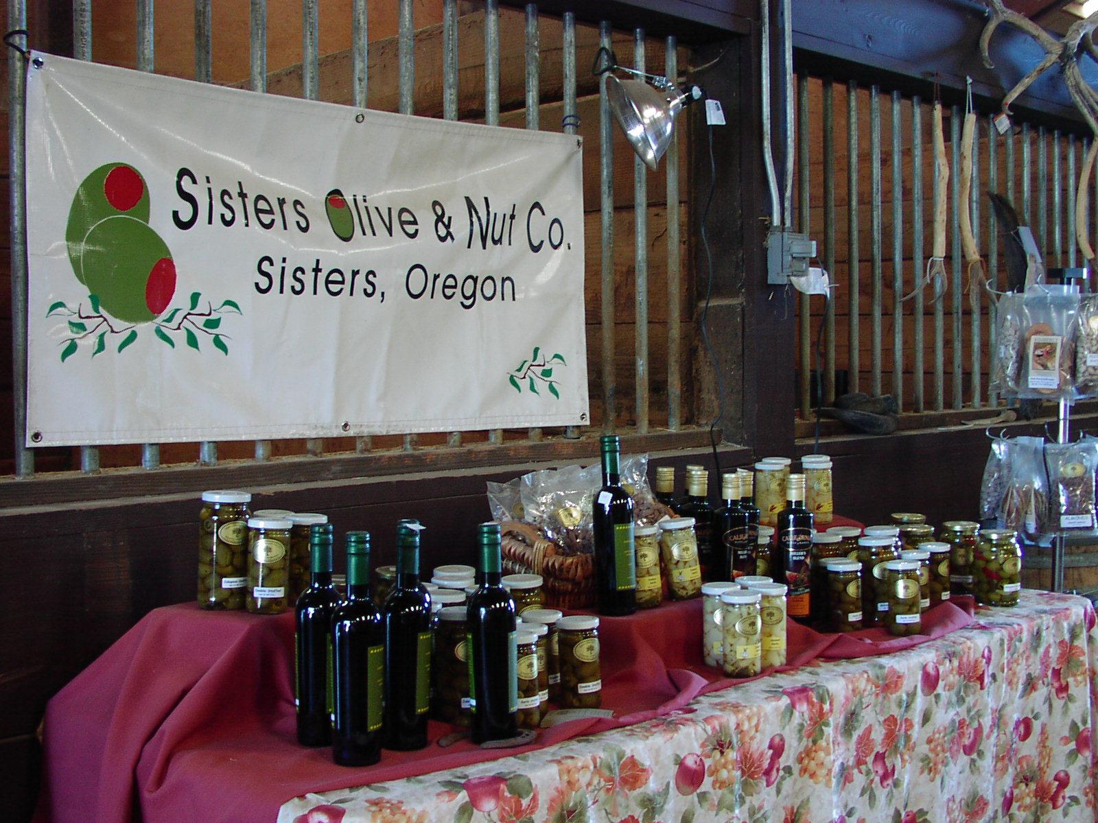 Market stall displaying bottles and jars of olive products under a banner.
