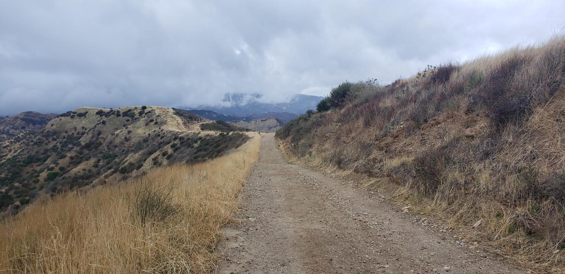 Dirt path through dry, grassy hills under a cloudy, overcast sky.