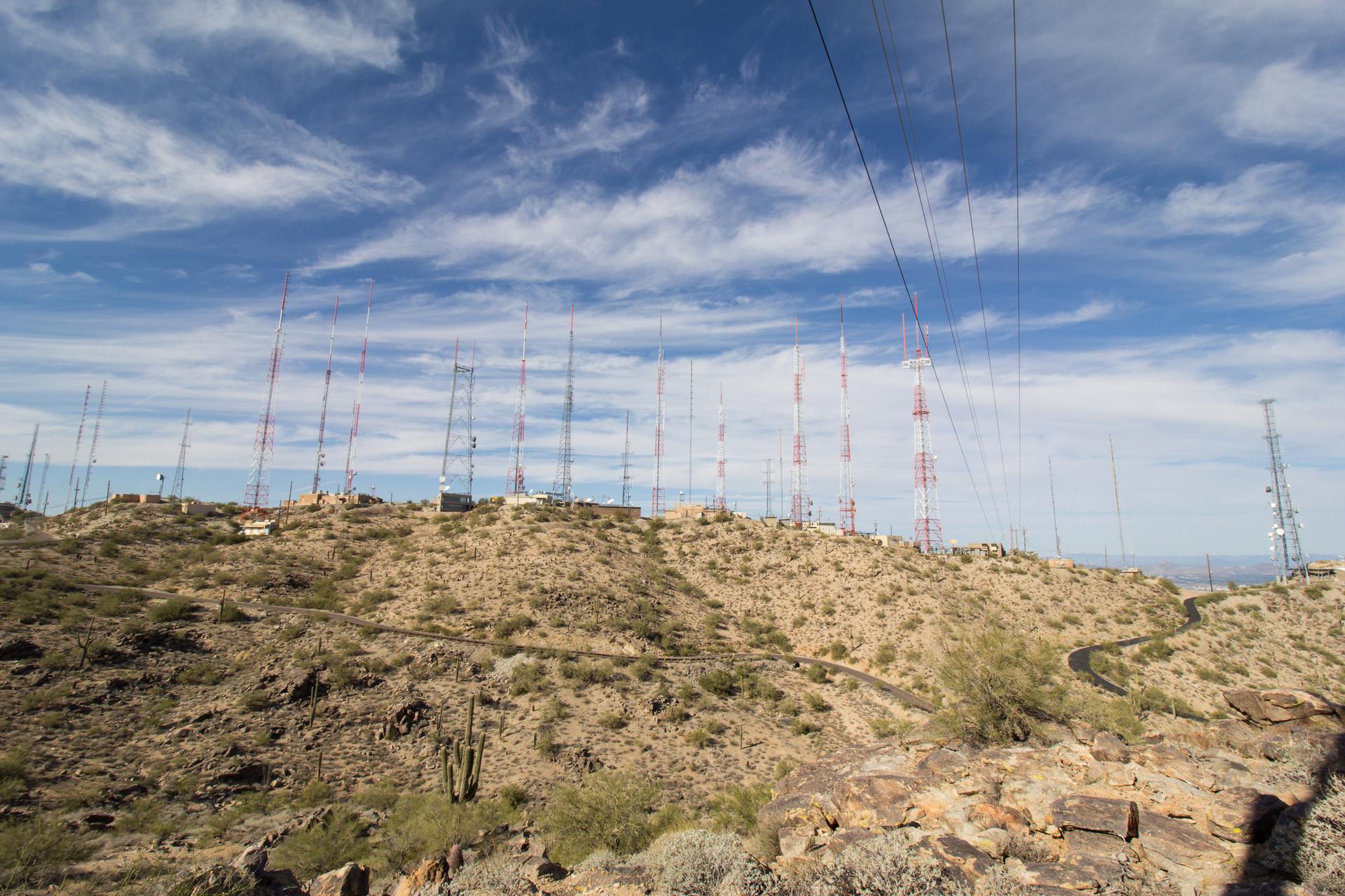 Numerous communication towers on a rocky hilltop under a cloudy blue sky.