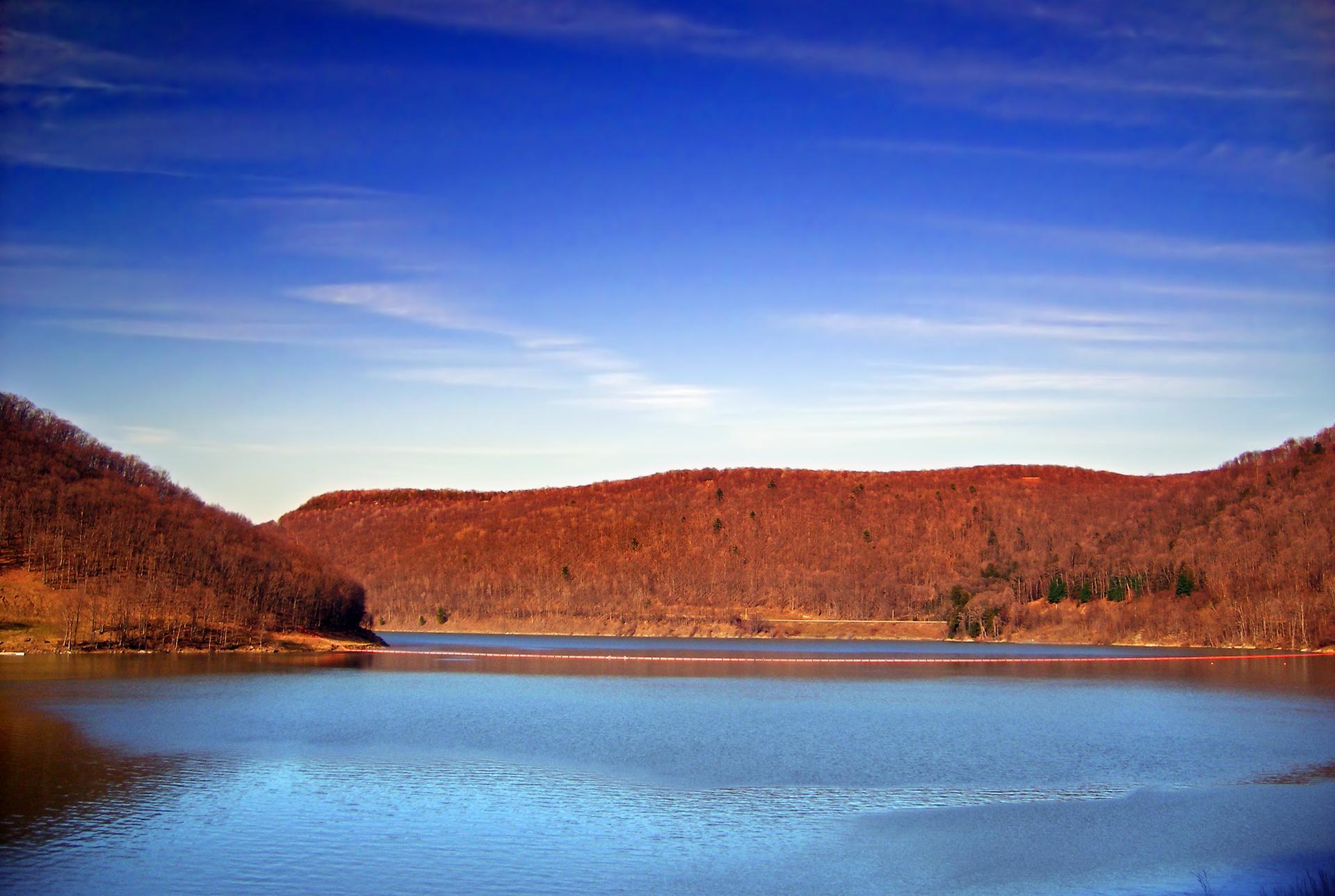 A wide blue lake surrounded by forested hills under a bright sky.