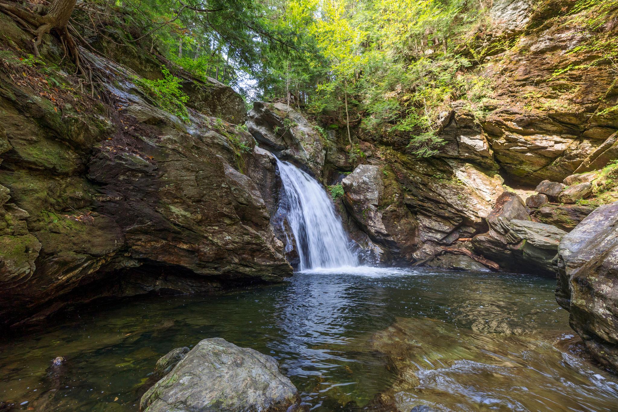 A waterfall cascades into a clear pool surrounded by rocky cliffs and trees.