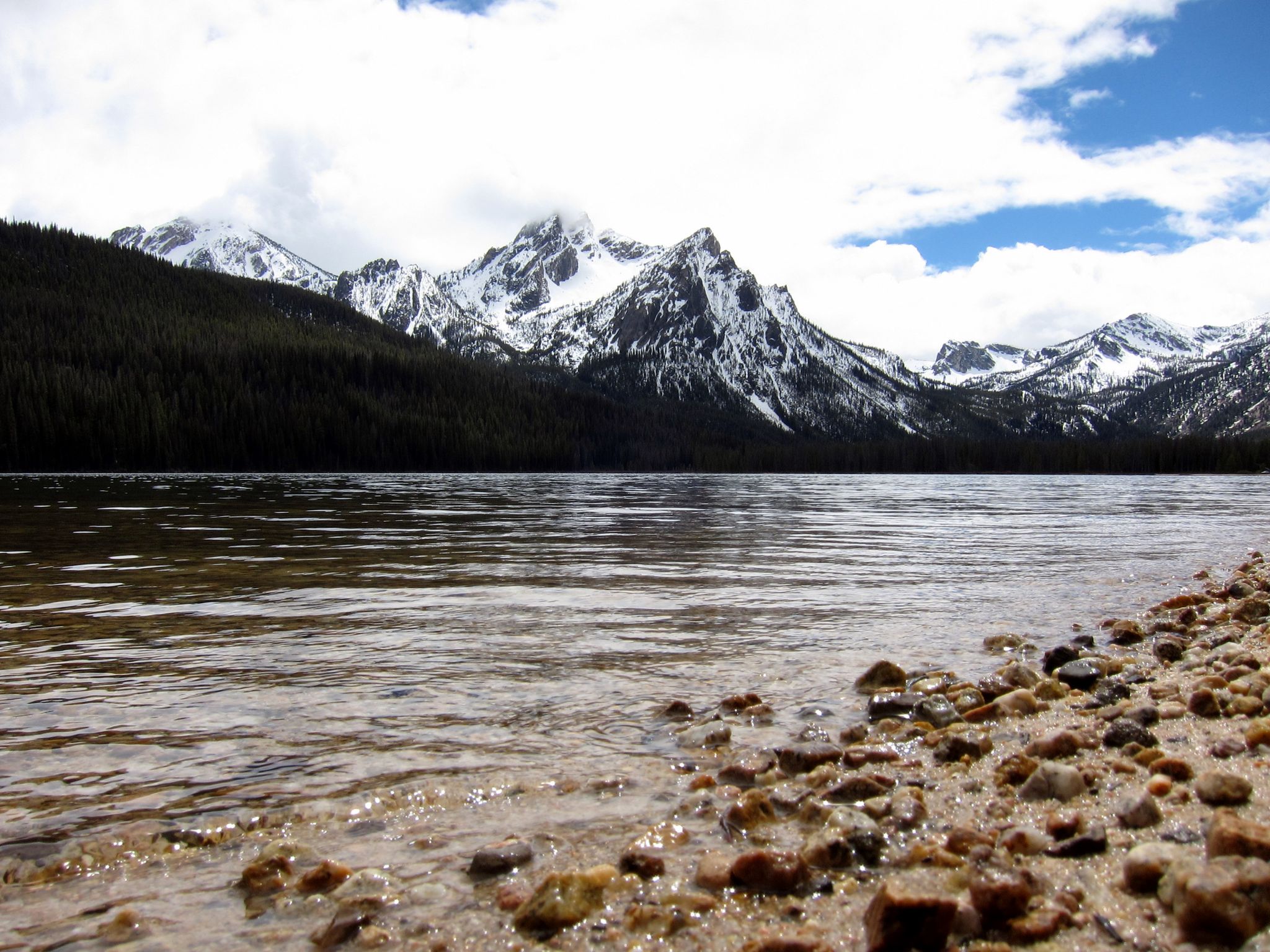 Rocky lake shore with snow-capped mountains and dark forests under a cloudy sky. - Stanley Lake loop from Stanley Lake