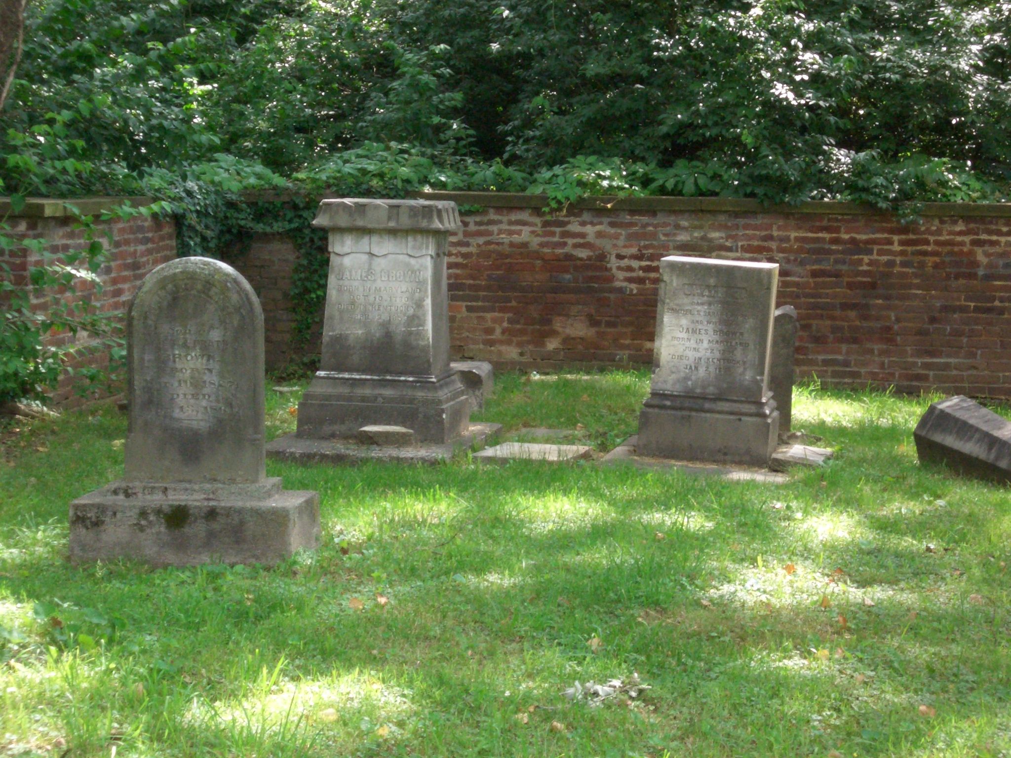 Several old gravestones in a green, sun-dappled field with a brick wall. - Brown Park Loop