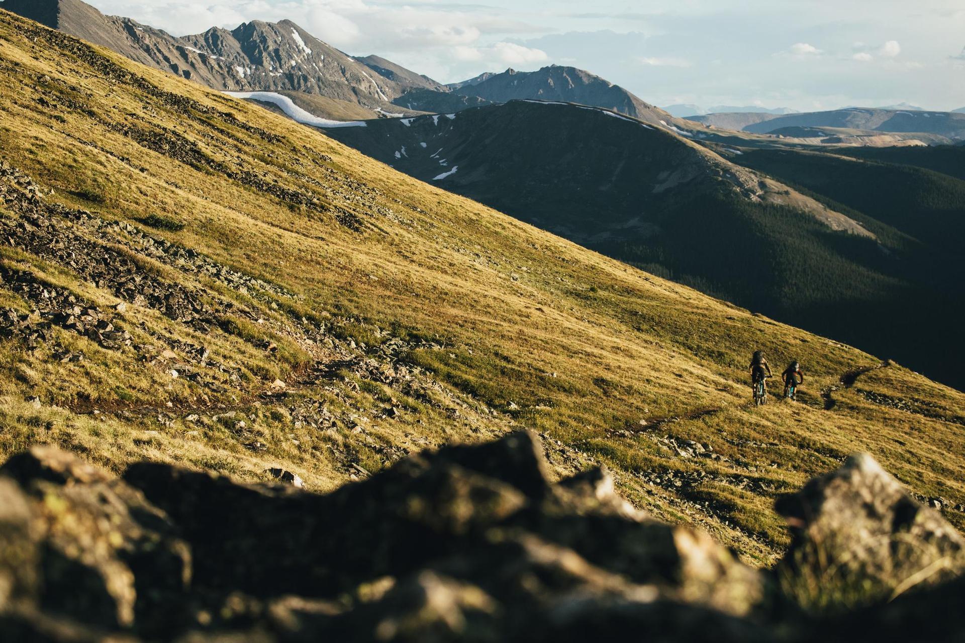 Two distant figures hike across a vast, golden mountain slope.