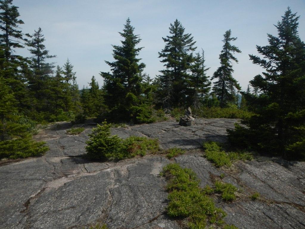 Rocky outcrop with evergreen trees and a small cairn under a bright sky.