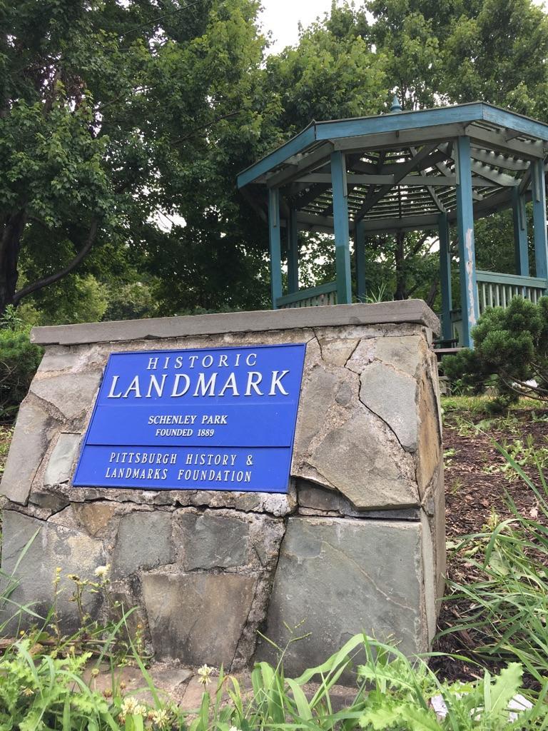 Stone sign with blue plaque in foreground, wooden gazebo and trees behind.
