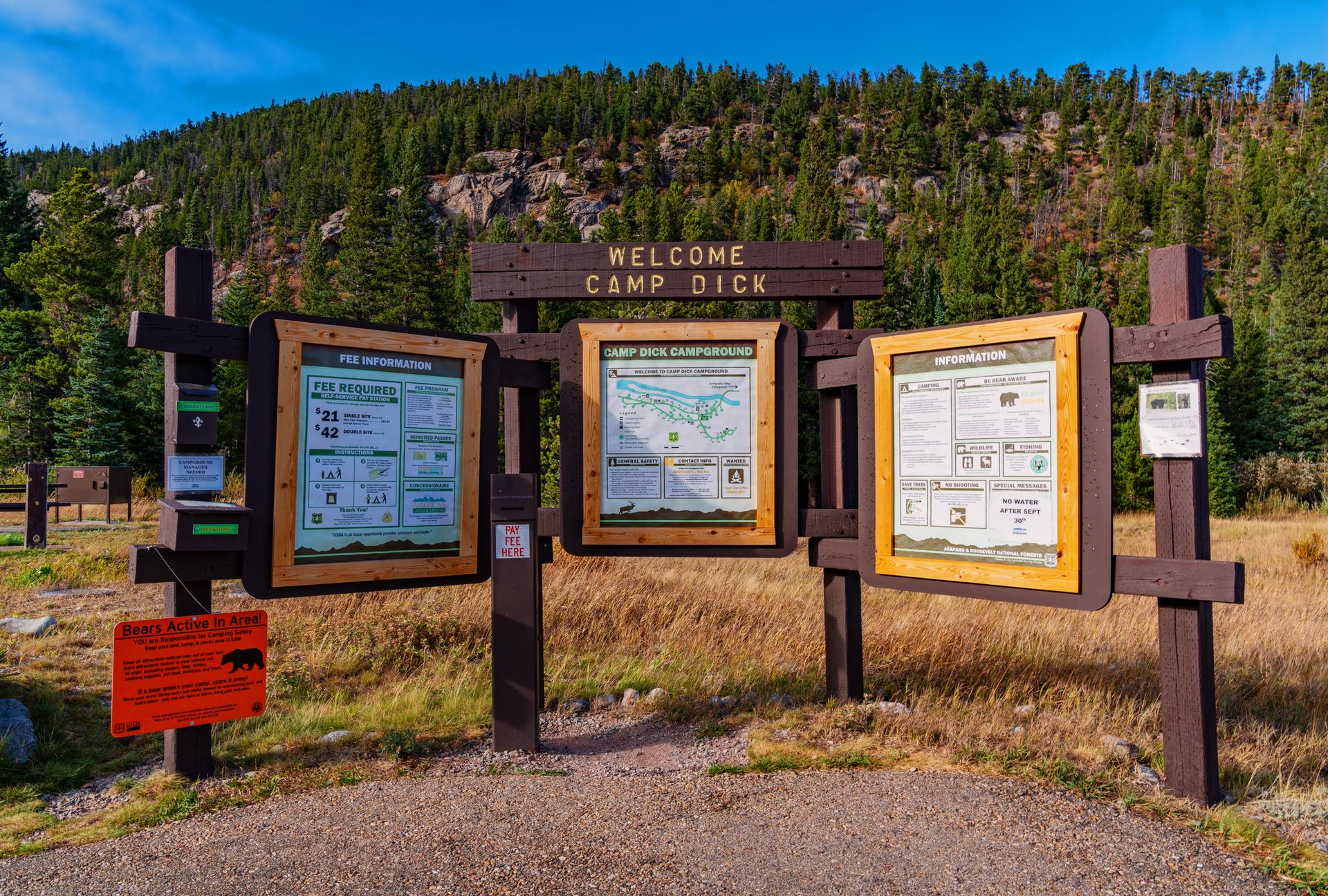 A wooden information kiosk with maps stands in a sunny, forested area.