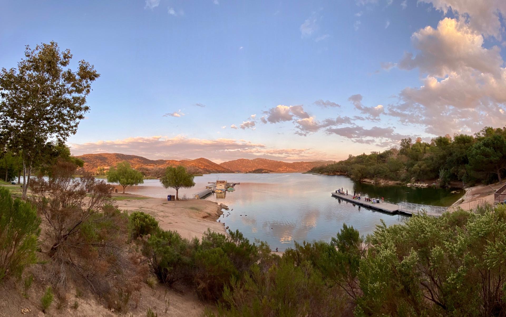 Panoramic lake view at sunset with distant hills and people on a floating dock.