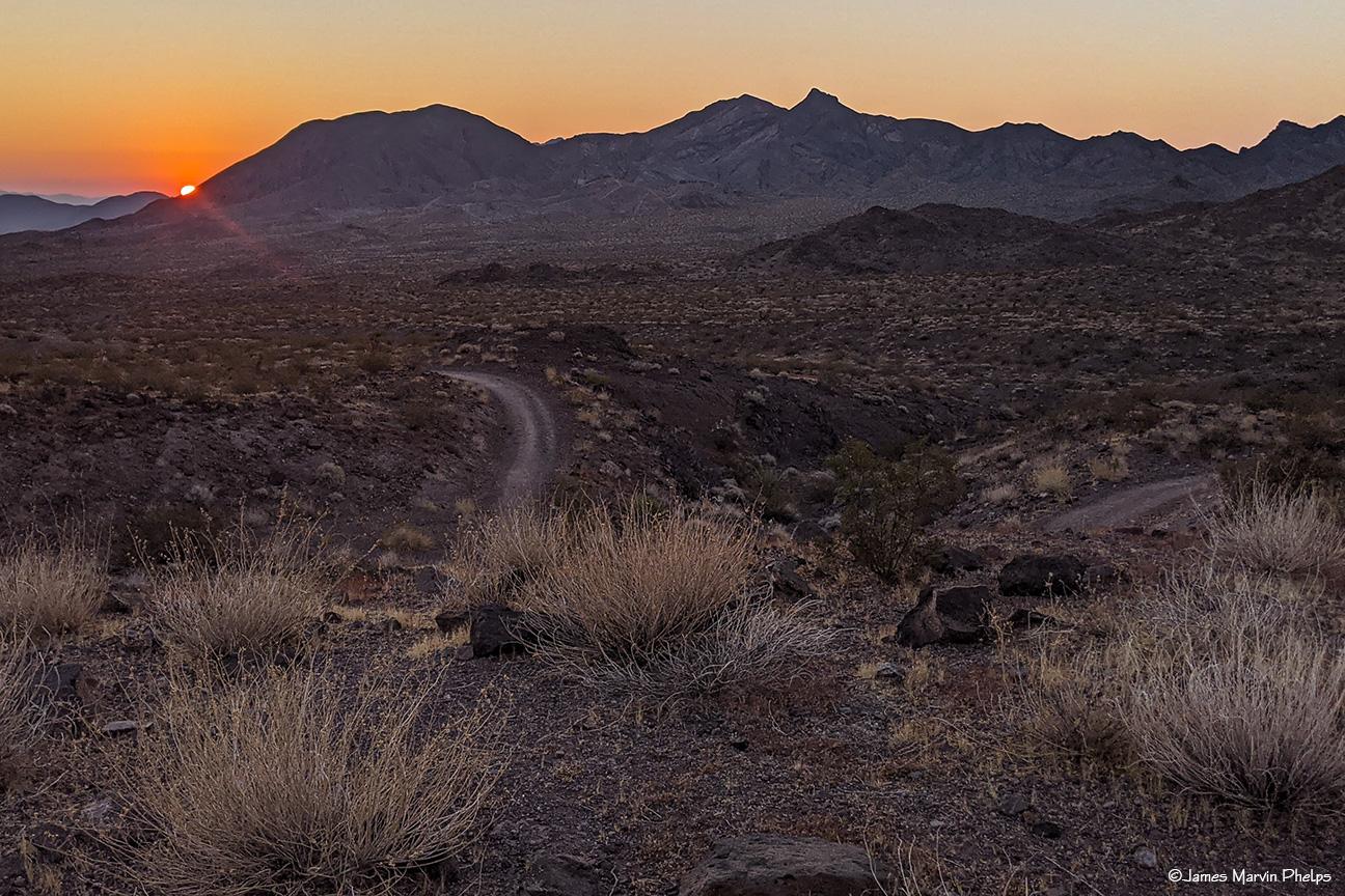 A desert landscape at sunset with a winding dirt road and distant mountains.