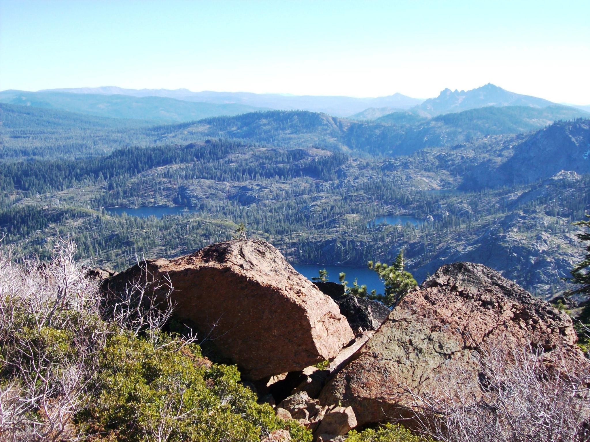 Vast mountain landscape with forested valleys, distant lakes, and foreground rocks.