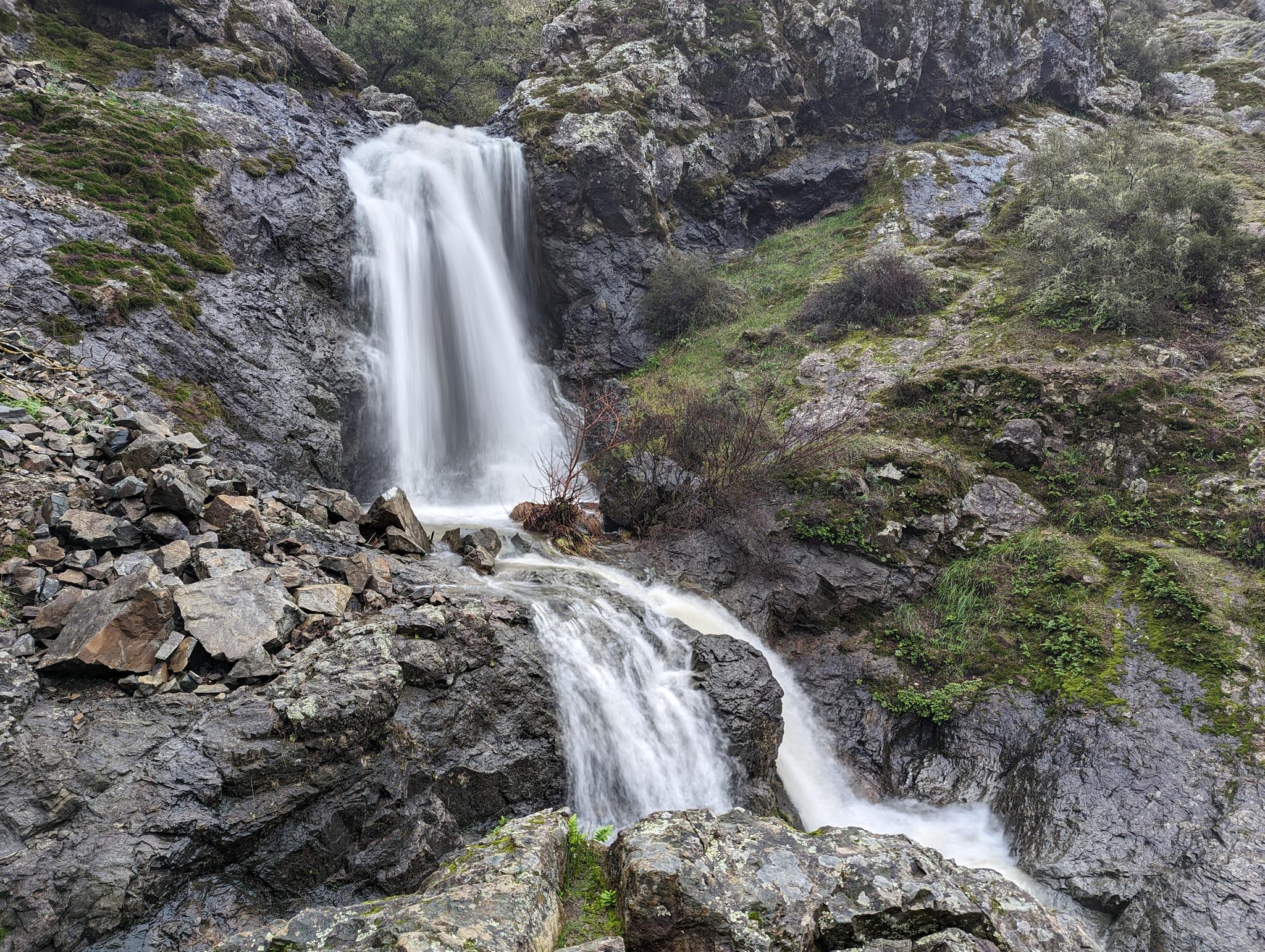 A powerful waterfall cascades down dark, mossy rocks on an overcast day.