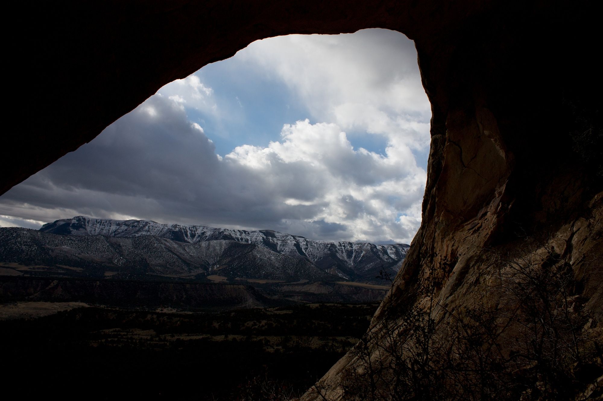 View from a dark cave opening of a cloudy sky and snow-capped mountains. - Rifle Arch Trail