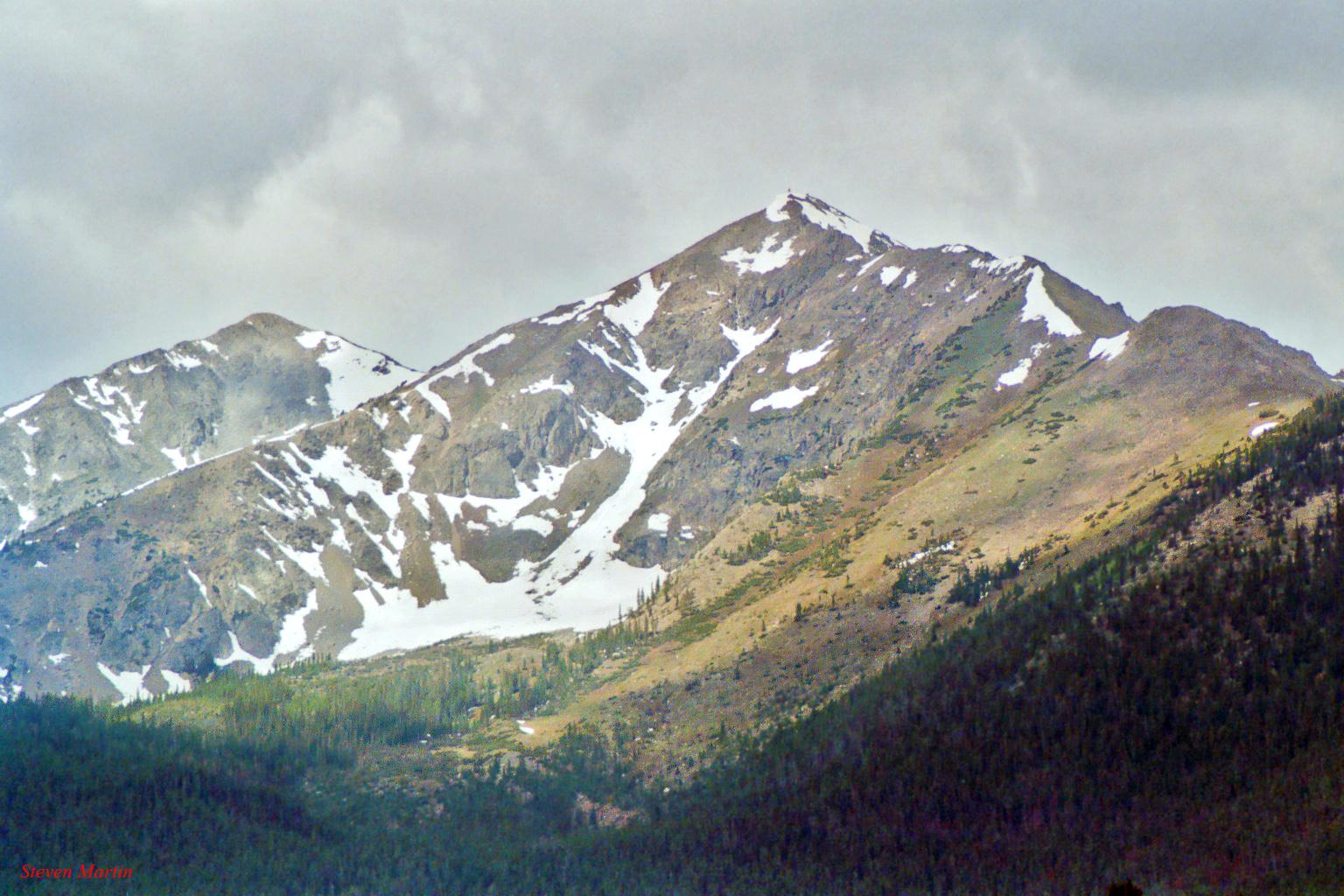 Rugged, snow-capped mountains under a cloudy sky, with forested lower slopes.