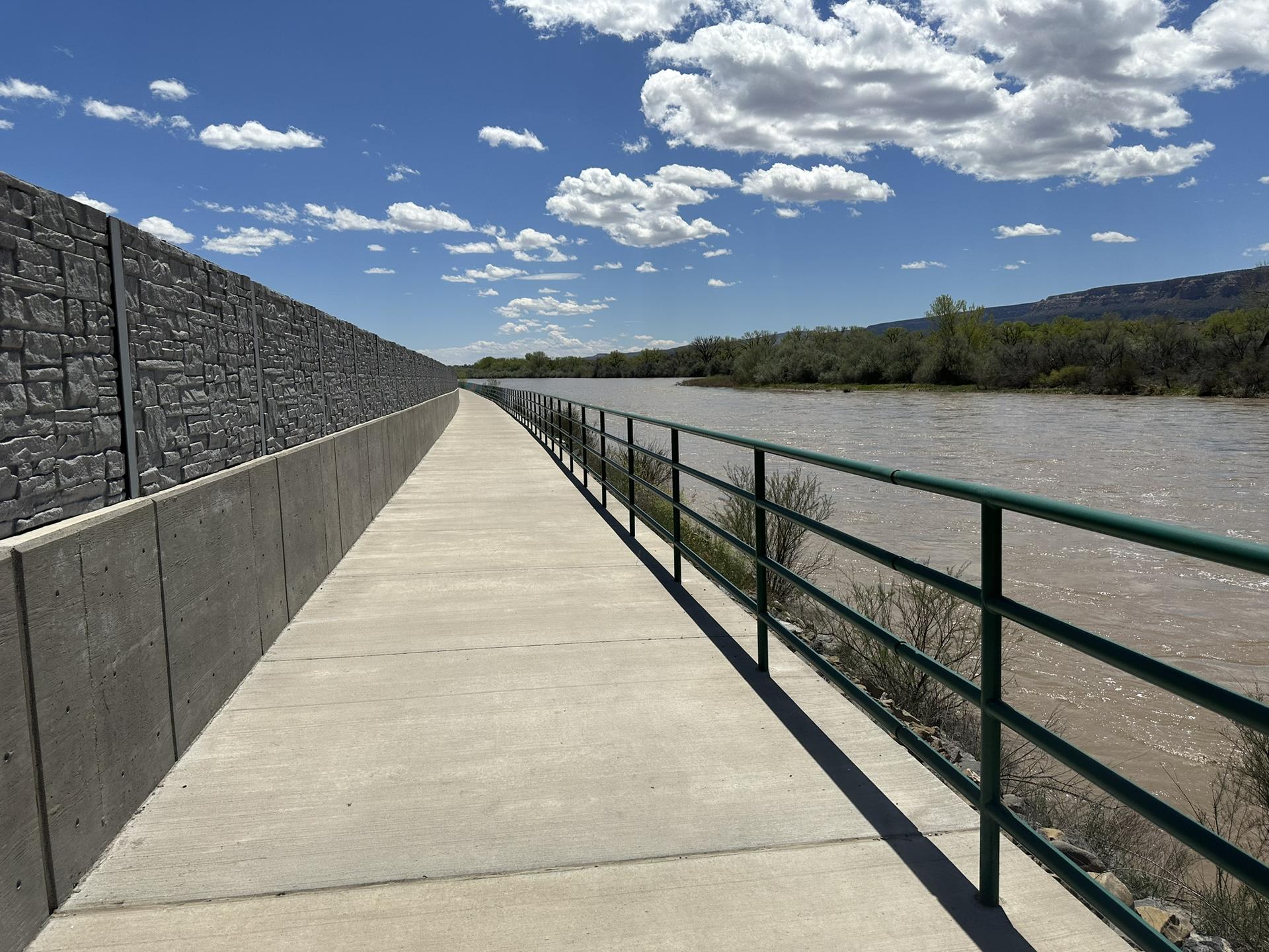 Paved path with green railing alongside a wide river under a cloudy sky.