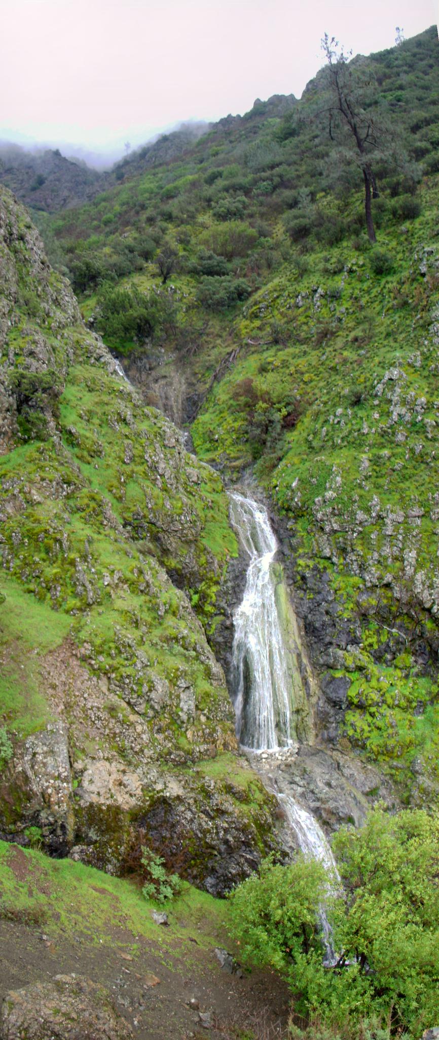 Multi-tiered waterfall cascading down a lush, green, rocky hillside on a cloudy day.