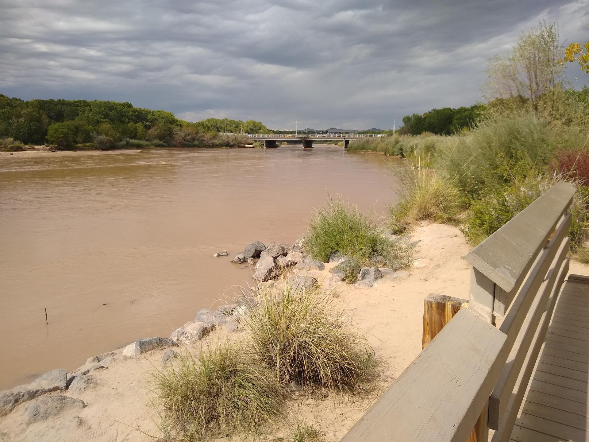 Muddy river with a distant bridge, sandy bank, and wooden railing.