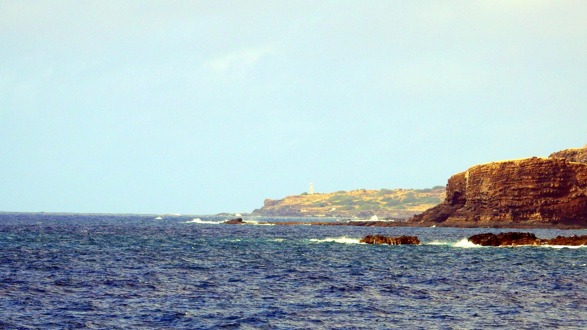 Choppy blue ocean meets brown cliffs under a hazy sky, distant structure visible.
