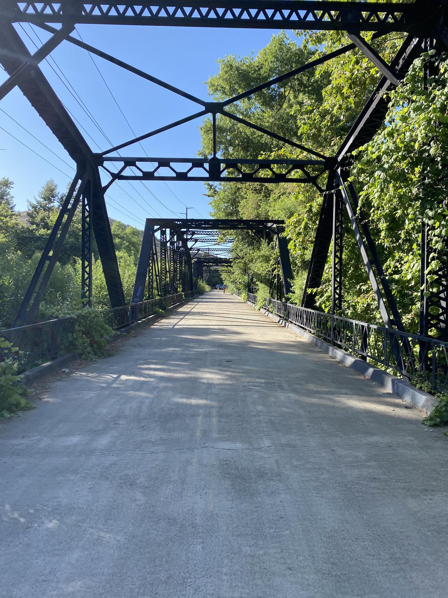 A long, black metal truss bridge spans a paved path under a clear sky.
