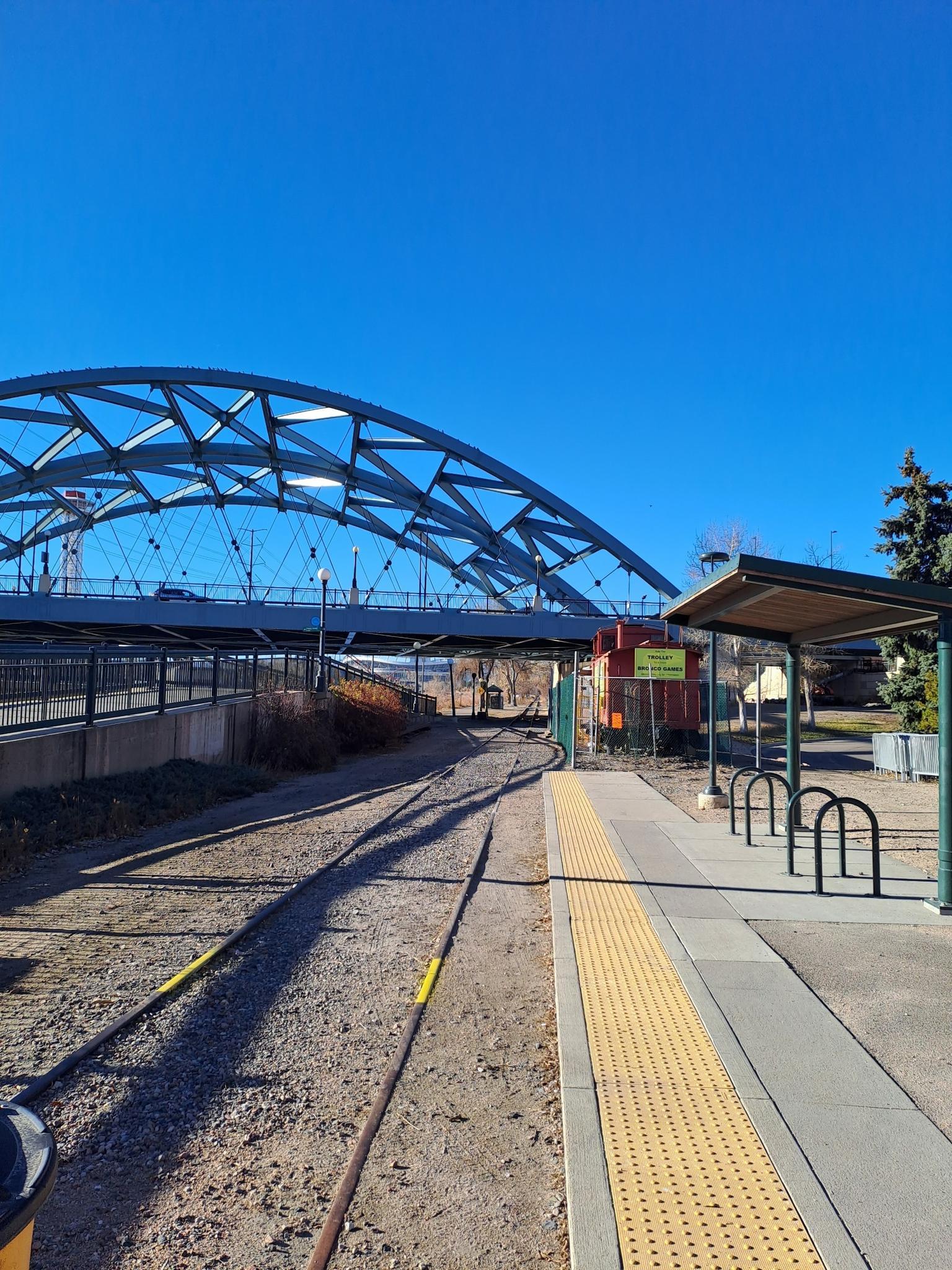 Train tracks and a platform lead towards a large arched bridge under a clear sky.