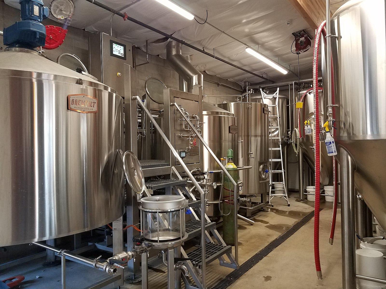 Brightly lit industrial room with large stainless steel brewing tanks and equipment.