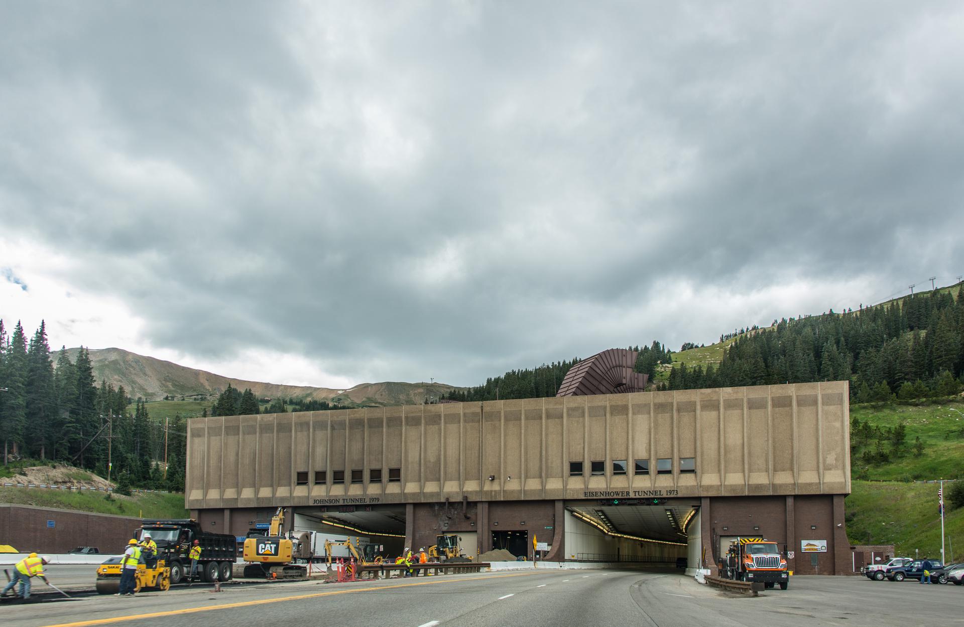 Construction workers and vehicles near a large tunnel entrance under an overcast sky.
