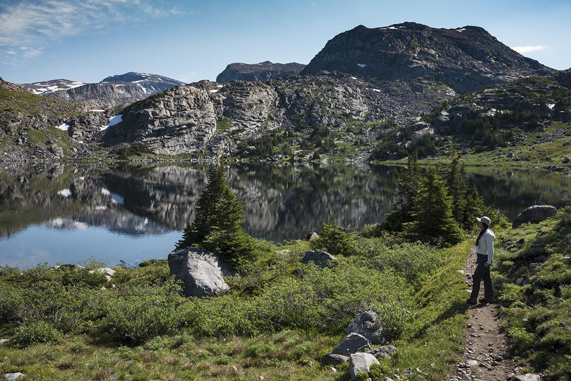 Hiker on a trail overlooking a serene mountain lake under sunny skies.