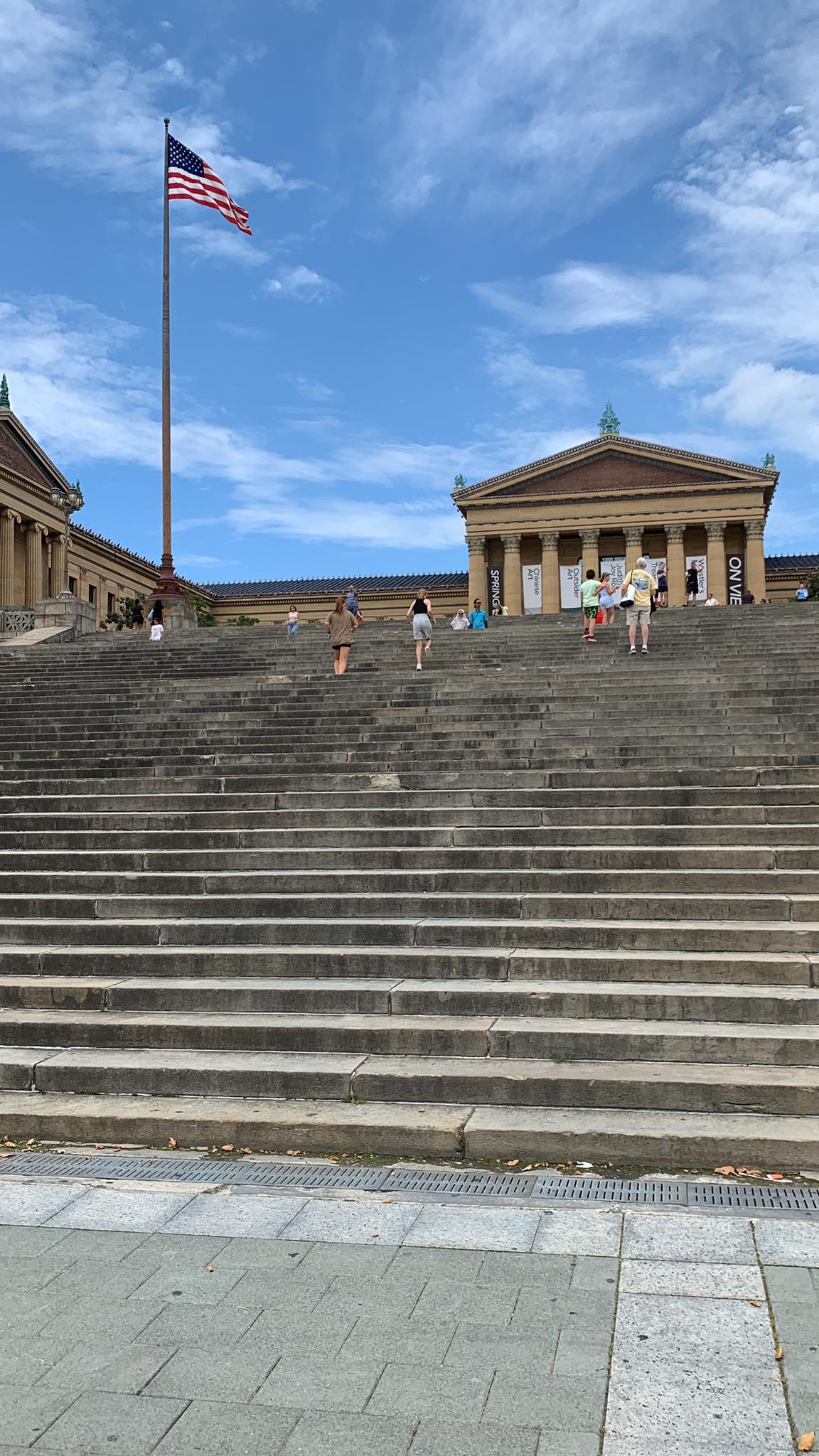 People ascend grand stone steps towards a classical building under a bright sky.