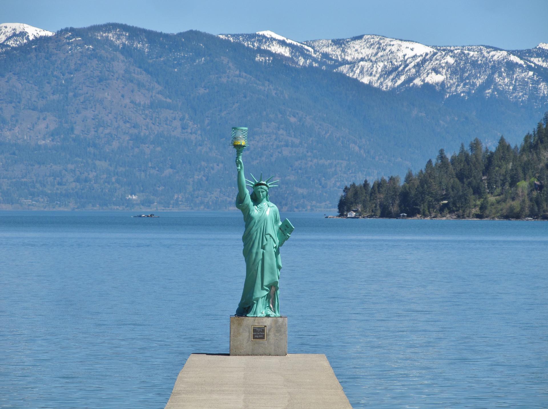 Statue of Liberty replica on a dock, calm water, forested mountains, bright day.
