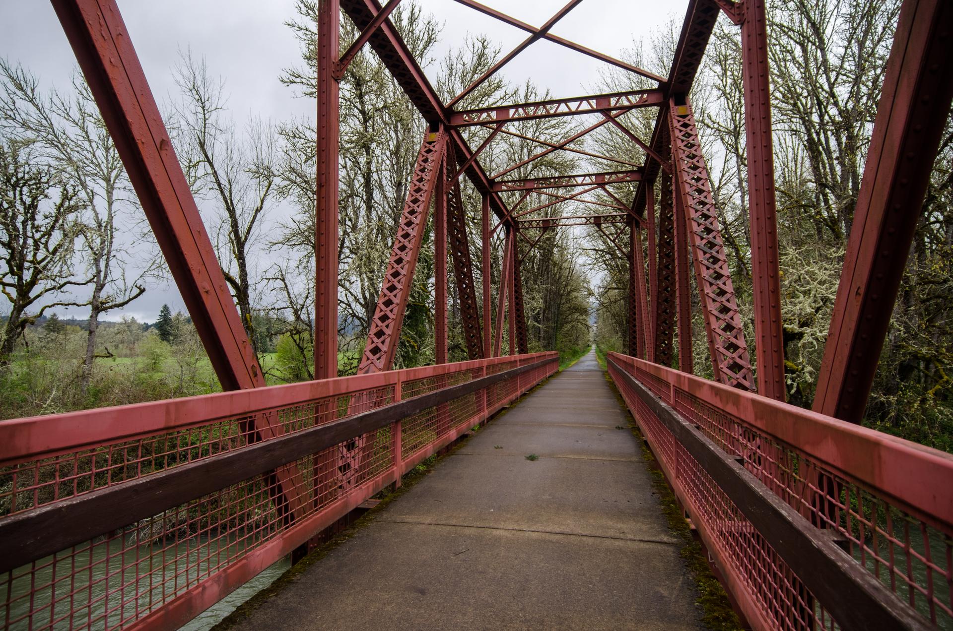 Empty red truss bridge pathway extends through trees on a cloudy day.