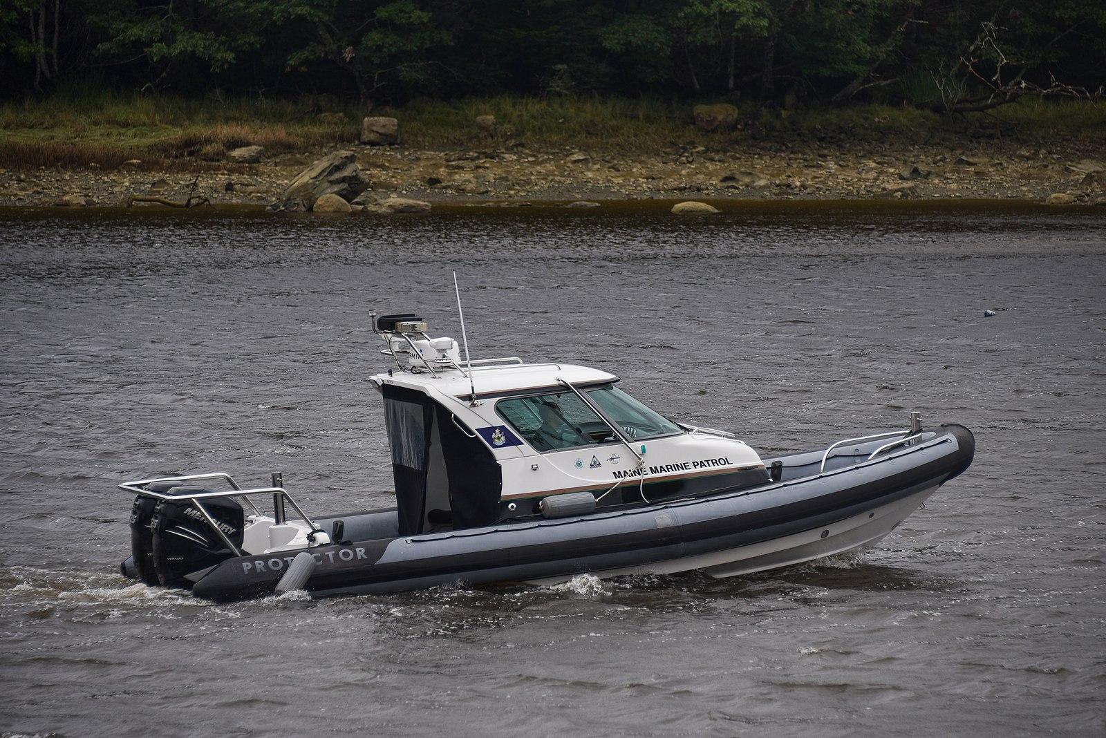 A white and grey patrol boat moves across dark water near a treeline.