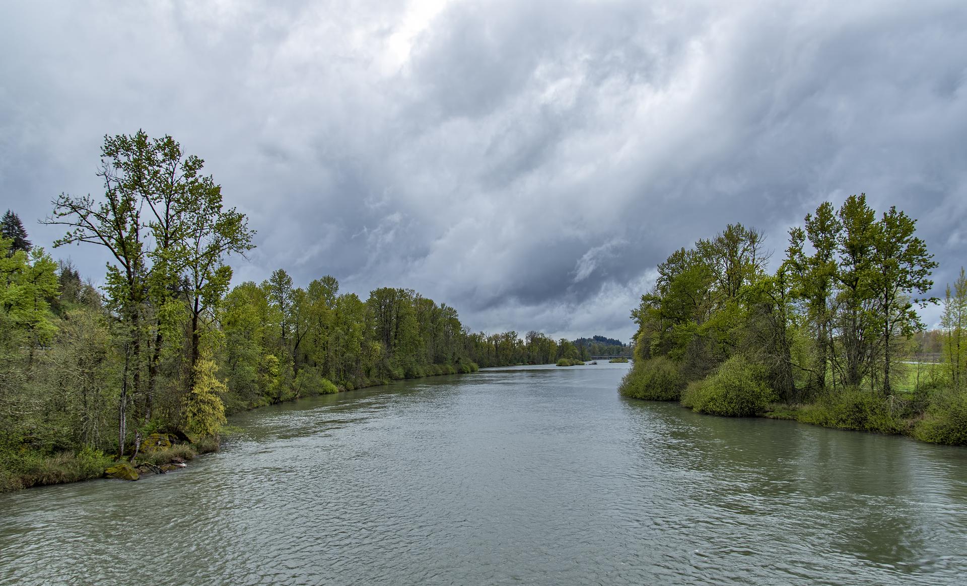 A wide river flows between lush green tree-lined banks under a cloudy sky.
